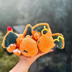 Könnte beinhalten: Ein handgefertigtes, orangefarbenes Charizard-Plüschtier aus Häkelarbeit mit grünen und gelben Akzenten. Das Plüschtier hat eine rote Spitze auf dem Kopf und weiße Akzente am Bauch. Das Spielzeug wird in einer Hand vor einem verschwommenen Hintergrund gehalten.