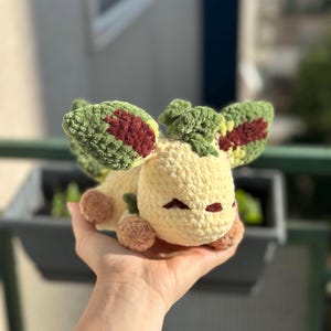 May include: A handmade crochet plush toy shaped like a creature with large, detailed ears. The toy is mainly cream-coloured with green and burgundy accents on the ears and brown accents on the feet. The plush is held in a hand.