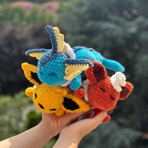 Peut inclure: Plusieurs peluches crochetées faites à la main, aux couleurs vives. On distingue des créatures jaunes, bleues et oranges, avec des détails blancs et noirs. Les jouets sont empilés.