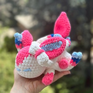 Peut inclure: Une peluche Sylveon faite à la main au crochet. Le jouet est composé de fil rose, blanc et bleu. Il a de grandes oreilles, un corps rond et de petits pieds. La peluche est tenue dans une main.