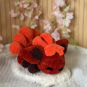 May include: A handmade crochet creature in shades of orange and red, with dark brown accents. The plush toy is resting on a fluffy white surface. The creature has a smiling face and is likely a decorative item.