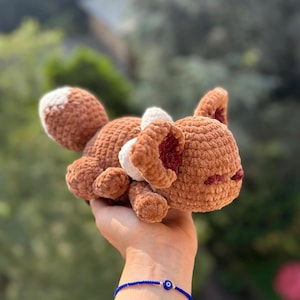 Può includere: Un peluche a forma di volpe realizzato all'uncinetto, nei toni del marrone, bianco e rosso. La volpe ha una consistenza soffice, con una macchia bianca sulla schiena e dettagli bianchi sulle orecchie. Il giocattolo è tenuto in mano.