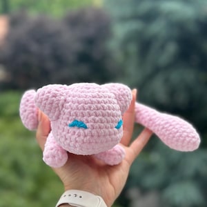 May include: A small, light pink crocheted plush toy featuring a rounded head, pointed ears, and a long, slender tail. The creature has embroidered bright blue, almond-shaped eyes that are closed, giving it a peaceful expression.