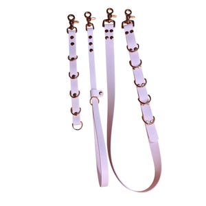 May include: Four white leather straps with gold-coloured hardware. The straps vary in length and design, including multiple gold rings and a handle. Each strap has a clasp at the top.