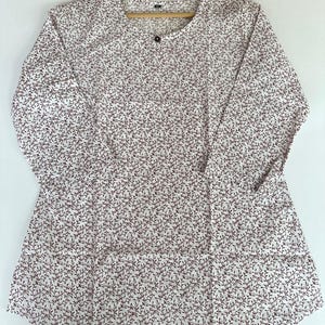 Pode incluir: Uma túnica branca de manga comprida com um padrão floral bordô. A túnica tem um decote redondo e um fecho de um botão. O tecido é leve, possivelmente algodão. A túnica está pendurada num cabide de madeira.