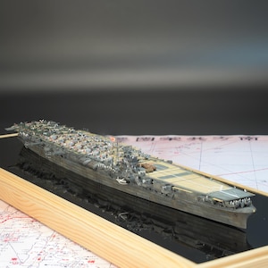 May include: A detailed model of a warship, displayed in a wooden frame. The model is gray and tan, with intricate details of the ship's structure. The frame sits on a map, with a black background.