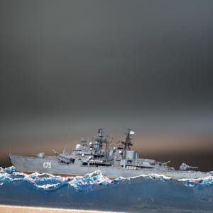 Puede incluir: Maqueta detallada de un buque de guerra gris, con la etiqueta "675", navegando sobre olas azules y blancas simuladas. El barco tiene múltiples torretas, antenas y una pequeña bandera blanca.