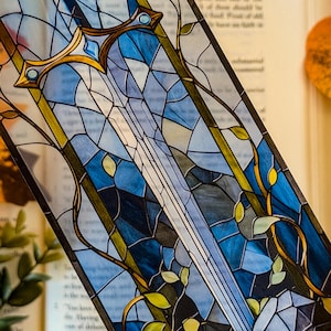 May include: A stained glass bookmark featuring a sword design. The bookmark has a blue and white color scheme with gold accents. Green leaves and vines are also incorporated into the design. The bookmark is placed on top of an open book.