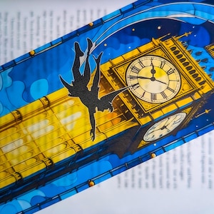 Puede incluir: Un marcapáginas con una ilustración estilizada del Big Ben y una silueta en vuelo. El diseño utiliza colores amarillo, azul y blanco, con un fondo de cielo estrellado. El marcapáginas está colocado en una página de libro.