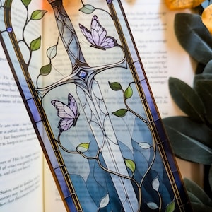 Può includere: Un segnalibro in vetro colorato con un disegno di spada, due farfalle lavanda e foglie verdi. Il segnalibro ha uno sfondo blu e trasparente, con un bordo scuro. È posto in un libro.