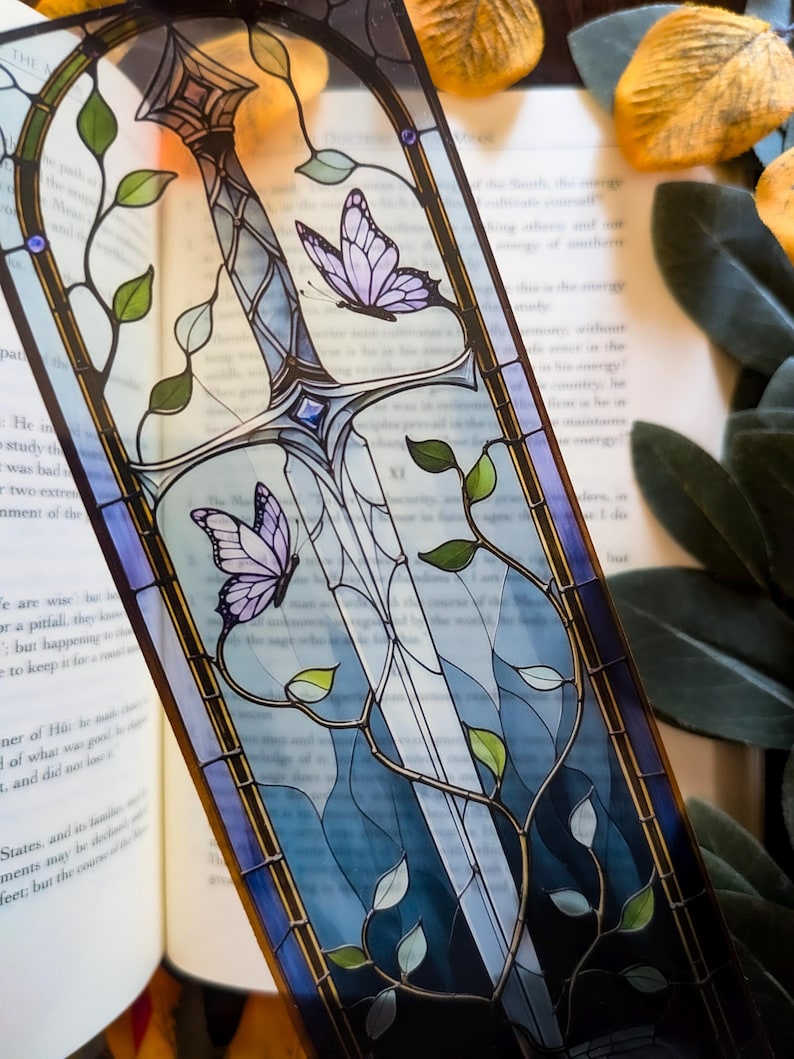 May include: A rectangular bookmark featuring a stained glass style illustration of a silver sword entwined with green vines and leaves. Two purple butterflies are positioned near the blade, and a small blue gem is set at the sword's crossguard.