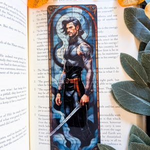 May include: A rectangular bookmark featuring a stained-glass style illustration of a dark-haired person with tattoos on their arms, wearing a black sleeveless outfit with an orange belt, and holding a silver sword against a blue background.