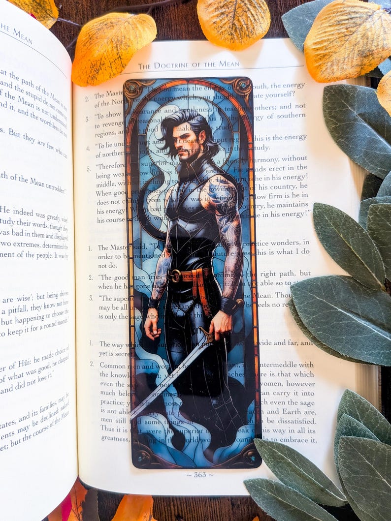 May include: A rectangular bookmark featuring a stained-glass style illustration of a dark-haired person with tattoos on their arms, wearing a black sleeveless outfit with an orange belt, and holding a silver sword against a blue background.