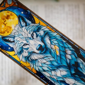 May include: A rectangular bookmark featuring a detailed illustration of a wolf with blue and white fur, set against a backdrop of a full yellow moon and starry night sky. The artwork has a stained-glass effect with a dark brown border.