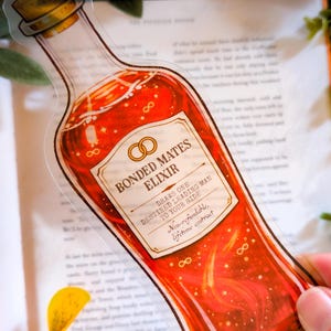 Puede incluir: Una pegatina transparente con forma de botella de poción, llena de líquido rojo y detalles dorados. La etiqueta dice "Bonded Mates Elixir" con texto adicional. La botella tiene un tapón de corcho.
