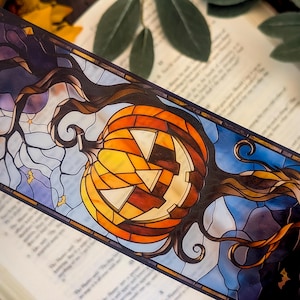 Op de afbeelding: Een rechthoekige boekenlegger met een illustratie in glas-in-loodstijl van een uitgesneden pompoen. Het ontwerp bevat donkere, gedraaide boomtakken, een maansikkel en kleine vleermuizen tegen een blauwe en paarse achtergrond.
