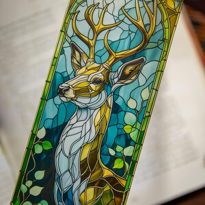 May include: A rectangular bookmark featuring a stained glass style illustration of a deer with large antlers. The design uses shades of blue, green, and gold, with intricate black outlines forming a mosaic pattern of leaves and geometric shapes.