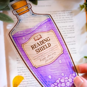 Puede incluir: Una pegatina transparente con forma de botella de poción con tapón de corcho. La botella contiene líquido morado y cristales blancos. La etiqueta dice "READING SHIELD" y "Deflects mid-chapter interrupters. Reapply after every plot-twist."
