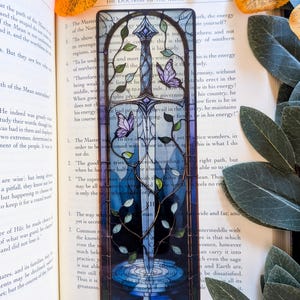 May include: A rectangular bookmark featuring a stained glass style illustration of a sword centered vertically. The sword is surrounded by climbing green vines and two purple butterflies. The background transitions from light blue to dark blue.