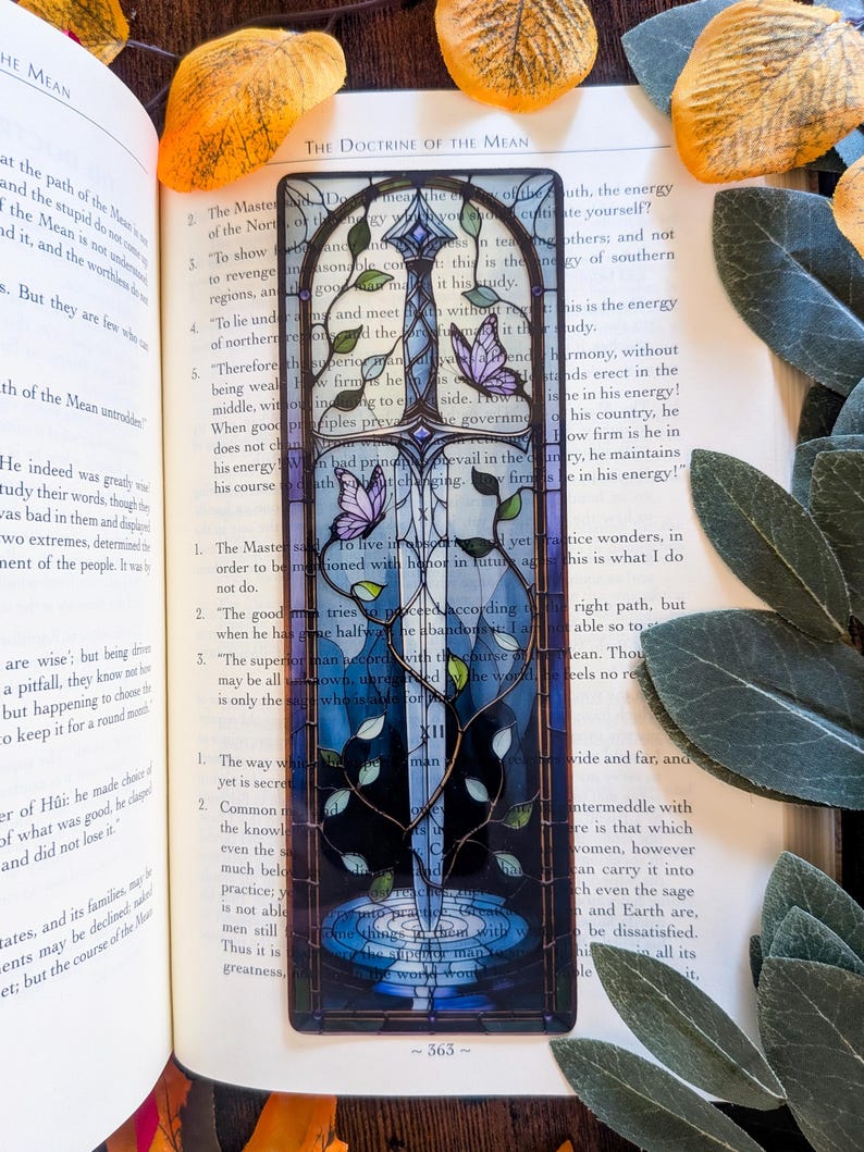 May include: A rectangular bookmark featuring a stained glass style illustration of a sword centered vertically. The sword is surrounded by climbing green vines and two purple butterflies. The background transitions from light blue to dark blue.