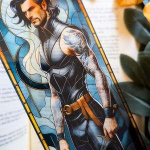 May include: A vertical bookmark featuring a stained-glass style illustration of a dark-haired individual with a beard, wearing a black sleeveless tunic, a brown belt, and arm cuffs. The figure holds a sword and has intricate tattoos on their arms.