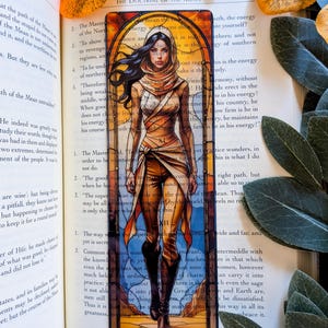 May include: A rectangular bookmark featuring a stained-glass style illustration of a person with long dark hair wearing a hooded tan outfit and brown boots. The artwork is framed by an arched border with orange, yellow, and blue glass panels.