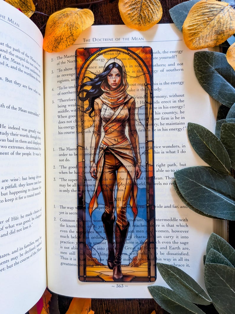 May include: A rectangular bookmark featuring a stained-glass style illustration of a person with long dark hair wearing a hooded tan outfit and brown boots. The artwork is framed by an arched border with orange, yellow, and blue glass panels.