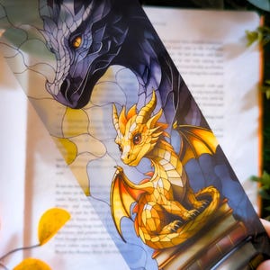 Puede incluir: Marcapáginas con una ilustración de vidriera de dos dragones. Un dragón dorado está sentado sobre libros, mientras que la cabeza de un dragón púrpura oscuro está arriba. El marcapáginas está sobre un libro abierto.