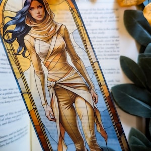 May include: A rectangular bookmark featuring a digital illustration of a person with long dark hair wearing a tan hooded outfit with wrapped fabric details. The character is framed by a yellow and brown stained glass window design.