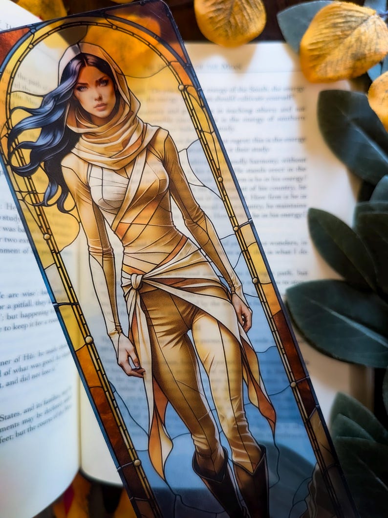 May include: A rectangular bookmark featuring a digital illustration of a person with long dark hair wearing a tan hooded outfit with wrapped fabric details. The character is framed by a yellow and brown stained glass window design.