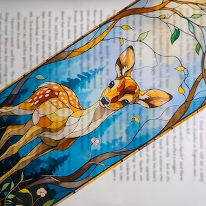May include: A decorative bookmark with a deer illustration. The deer is rendered in orange, yellow, and brown, set against a blue backdrop with tree branches. The bookmark is rectangular and seems to be made of a translucent material.