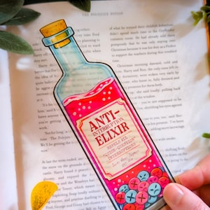 Puede incluir: Una pegatina transparente con forma de botella de poción, con la etiqueta "Anti-Interruption Elixir". La etiqueta rosa y roja dice "Repels all quick-question interruptions". La pegatina tiene una tapa de corcho y un líquido rojo.