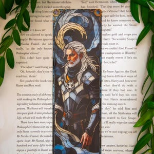 May include: A bookmark with a stained glass effect, depicting a man with a long white beard and hair, in armour. The design uses blue, white, and gold, with a wolf emblem. The bookmark is on a book page.