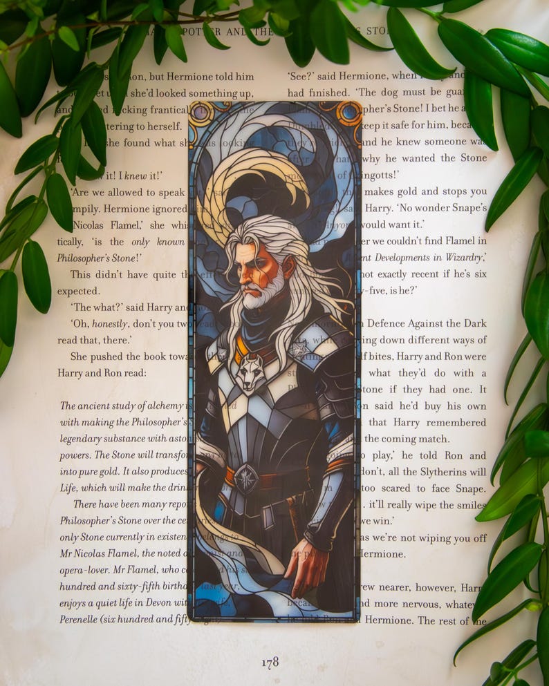 May include: A bookmark with a stained glass effect, depicting a man with a long white beard and hair, in armour. The design uses blue, white, and gold, with a wolf emblem. The bookmark is on a book page.