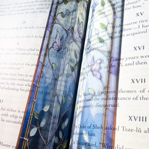 May include: A translucent bookmark featuring a stained glass design with blue and purple hues, intricate black lead lines, green leaves, and purple butterflies. The bookmark is placed between the pages of an open book with printed text.