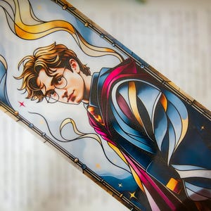 Könnte beinhalten: Rechteckiger Lesezeichen mit einer Illustration im Buntglasstil eines jungen Mannes mit Brille. Das Design verwendet Blau-, Gold- und Rosatöne mit Sternendetails. Das Lesezeichen befindet sich vor einem verschwommenen Hintergrund.