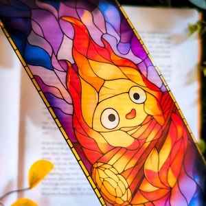 Puede incluir: Un marcapáginas rectangular con una ilustración estilo vidriera de un personaje de fuego de dibujos animados con ojos grandes y boca pequeña. El diseño utiliza tonos vibrantes de llamas naranjas, amarillas y rojas sobre un fondo morado y azul.