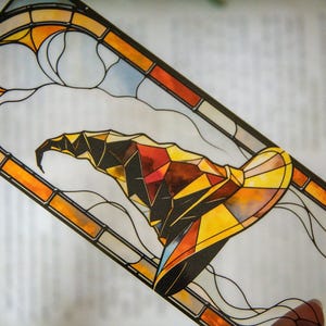 May include: A stained glass bookmark featuring a colorful, geometric wizard's hat with a pointed tip. The hat is set against a background of yellow, orange, and red stained glass.