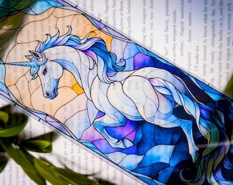 Puede incluir: Un marcapáginas de vidriera con un unicornio blanco con melena y cuerno azules. El diseño incorpora tonos de azul, morado y blanco, con un fondo de tonos cálidos. El marcapáginas es rectangular y parece ser de un material transparente.