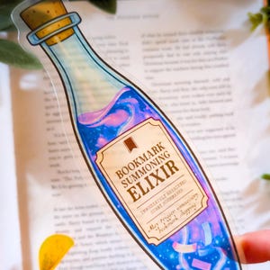 Puede incluir: Un marcapáginas transparente con forma de botella de poción, lleno de líquido azul y rosa arremolinado. La etiqueta dice "Bookmark Summoning Elixir" con texto adicional. El marcapáginas está sobre un libro abierto.