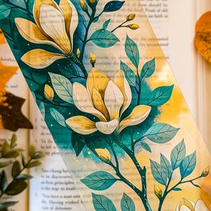 May include: A rectangular transparent bookmark featuring a botanical illustration of cream-coloured magnolia flowers with yellow centres, dark teal leaves, and thin branches. The design includes small gold speckles scattered across the surface.