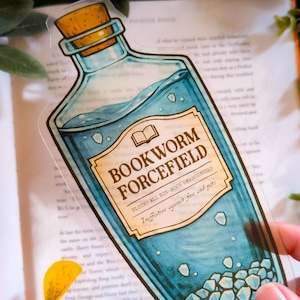 Puede incluir: Una pegatina transparente con forma de botella de poción, con tapa de corcho y líquido azul. La etiqueta dice "BOOKWORM FORCEFIELD" y "BLOCKS ALL NON-BOOK EMERGENCIES". La pegatina tiene forma de botella de poción.