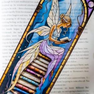 May include: A rectangular bookmark featuring a stained glass style illustration of a fairy with translucent wings sitting on a stack of colorful books. The fairy has blonde hair and wears a light blue and purple dress while reading a small book.