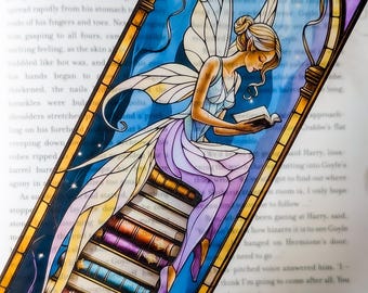 Puede incluir: Un marcapáginas estilo vidriera que representa a un hada leyendo un libro. El hada tiene cabello rubio, alas y viste un vestido azul claro y morado. El marcapáginas tiene un fondo azul con estrellas y una pila de libros.