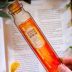 Puede incluir: Un marcapáginas transparente con forma de botella, con una tapa de corcho y una etiqueta que dice "Deadline Survival Serum". La etiqueta también indica "Unlocks Emergency Focus Mode" y "May reduce politeness". El líquido interior es naranja.