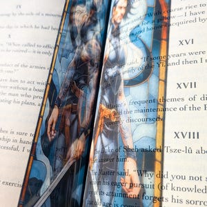 May include: A vertical bookmark featuring a stained-glass style illustration of a person with dark hair and a sword, set against a blue and orange background. The bookmark is placed between the pages of an open book with visible printed text.