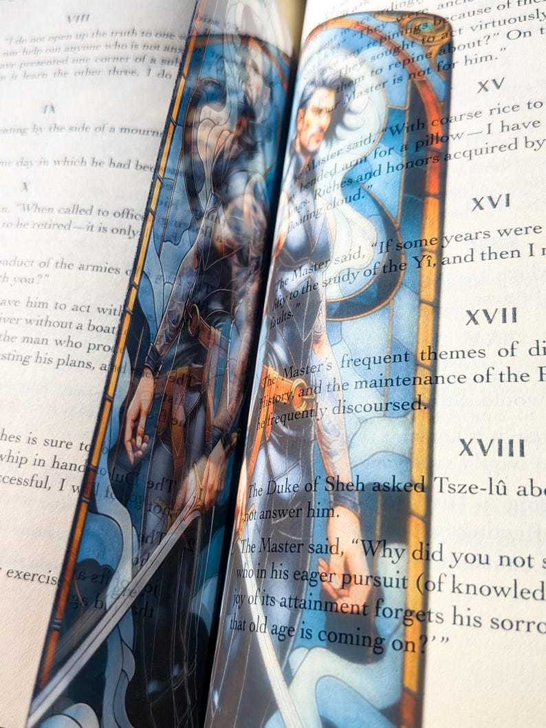 May include: A vertical bookmark featuring a stained-glass style illustration of a person with dark hair and a sword, set against a blue and orange background. The bookmark is placed between the pages of an open book with visible printed text.