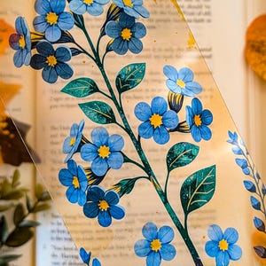 May include: A clear, rectangular bookmark featuring a detailed illustration of blue forget-me-not flowers with yellow centres and green leaves. The design includes subtle gold-coloured accents and speckles scattered across the transparent surface.
