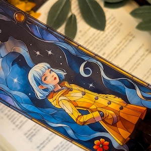 May include: A rectangular bookmark featuring a stained glass style illustration of a person with short white hair wearing a yellow coat. A black cat silhouette is visible in the upper left corner against a dark blue starry sky with swirling blue clouds.