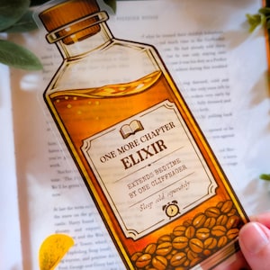 Puede incluir: Una pegatina transparente con forma de botella de poción, llena de líquido ámbar y granos de café. La etiqueta dice "ONE MORE CHAPTER ELIXIR" y "EXTENDS BEDTIME BY ONE CLIFFNAGER".